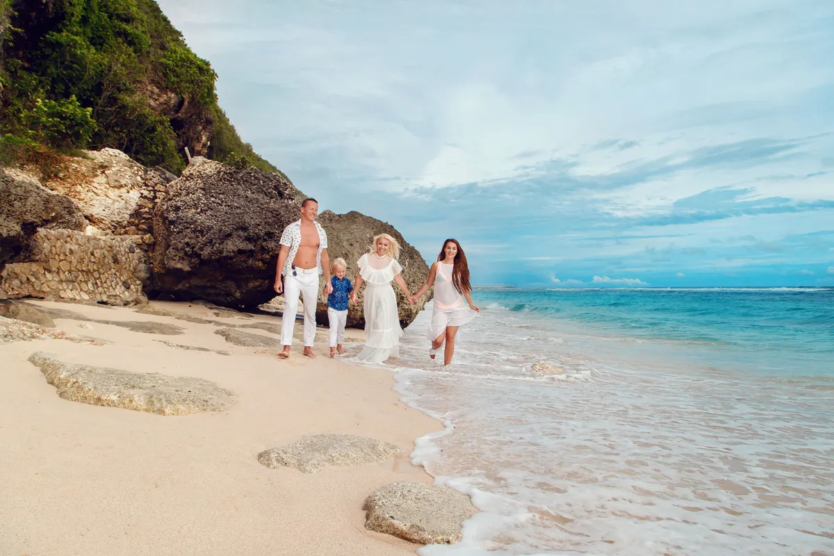A family walking together along a white sand beach with turquoise water and cliffs in Bali, Indonesia
